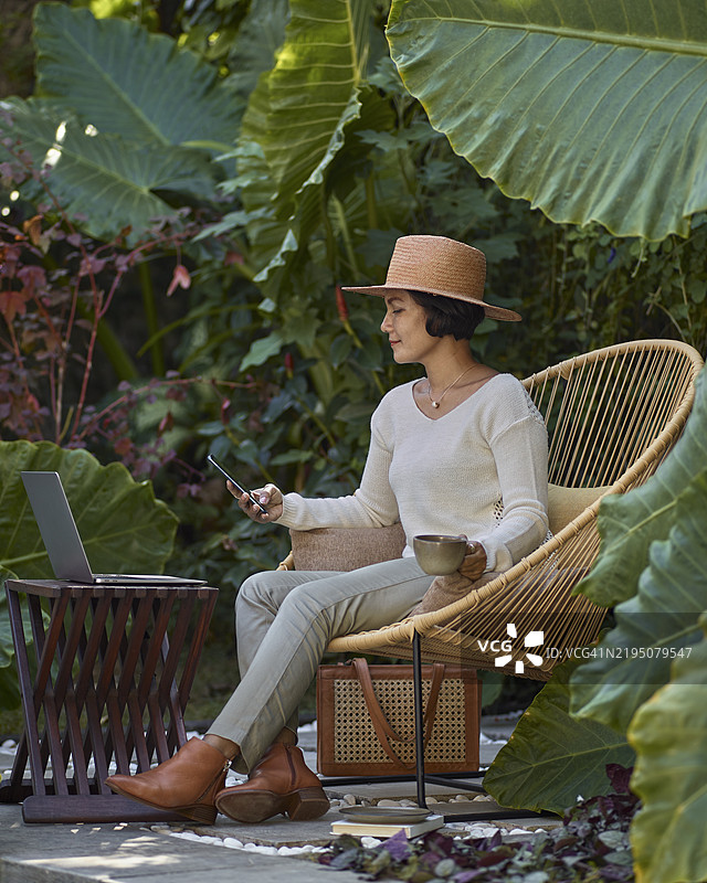 亚洲女性在家工作，坐在花园家具上，周围环绕着热带植物，使用手机。图片素材