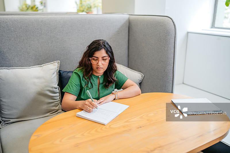 中年印度女性在澳大利亚悉尼的办公大楼内的桌子上写笔记。图片素材