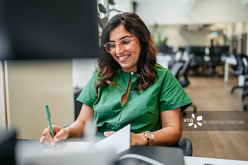 中年印度女性在澳大利亚悉尼的办公大楼内，一边在电脑上工作，一边在笔记本上写字。图片素材