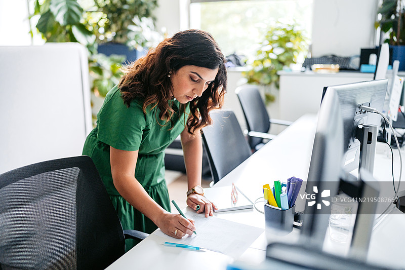 中年印度女性在澳大利亚悉尼的办公大楼里，一边在电脑上工作，一边在笔记本上写字。图片素材
