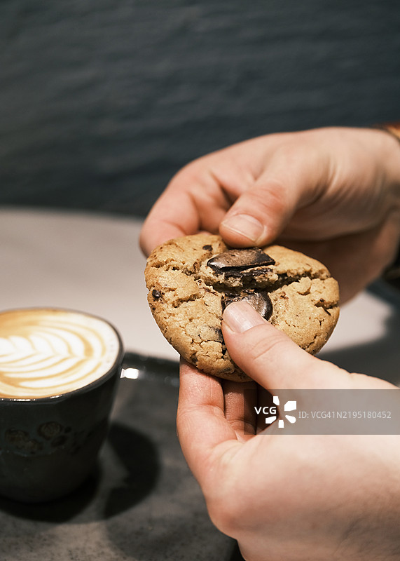 手握刚出炉的巧克力曲奇饼干，饼干中心流出融化的巧克力，旁边是一杯泡沫丰富的拿铁，营造出温馨舒适的咖啡馆氛围，奢华的甜点与咖啡搭配概念。图片素材