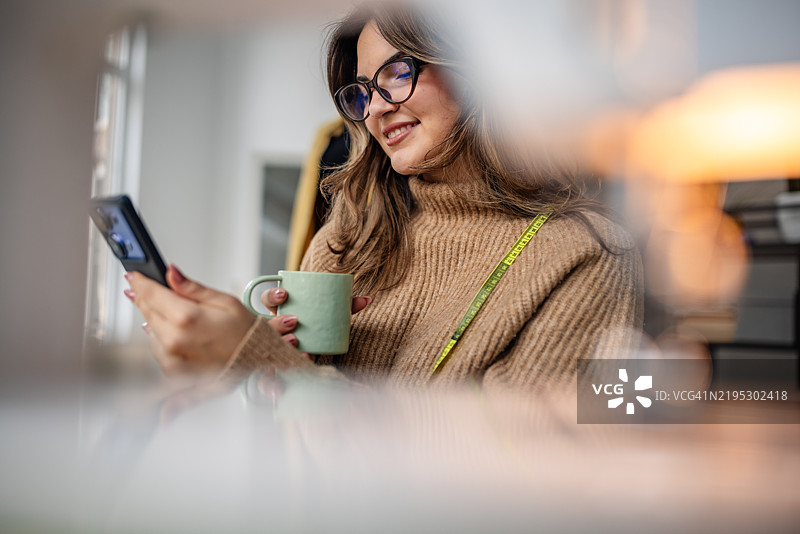 微笑的女性穿着舒适的毛衣，在光线充足的室内空间里享用饮品，同时使用手机。图片素材