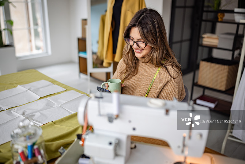 女人在温馨明亮的工作室里享受热饮，同时进行缝纫项目。图片素材