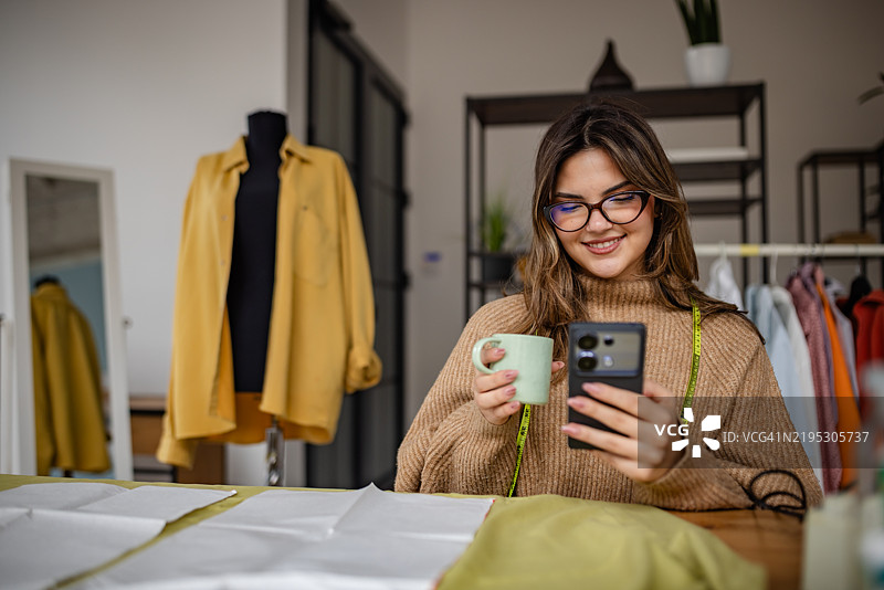 穿着舒适毛衣的女性在明亮的服装设计工作室中享受咖啡，同时使用智能手机。图片素材