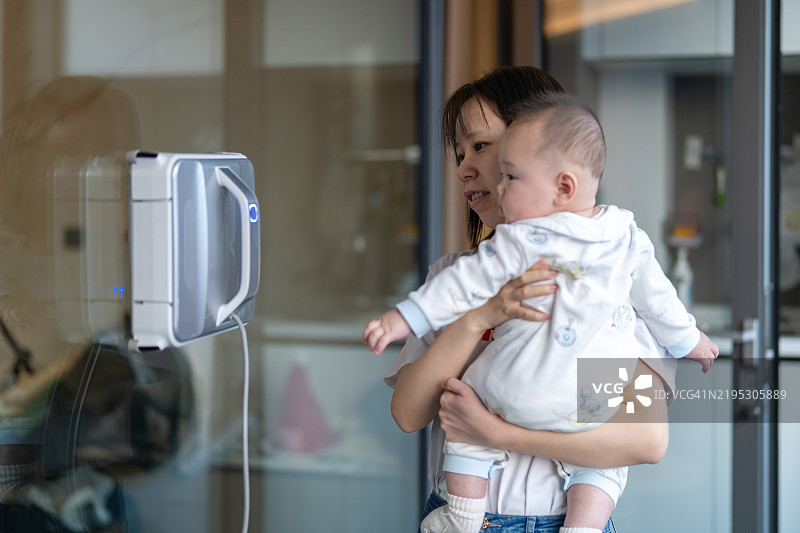 一位亚洲妈妈抱着她的宝宝，正在看着窗户清洁机器人工作。图片素材