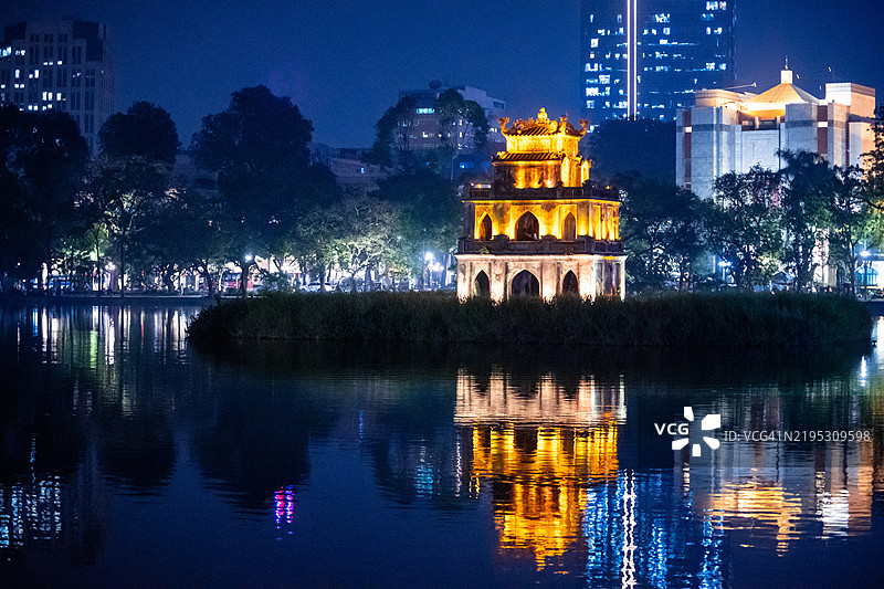夜晚照亮的传统风格的龟塔倒映在还剑湖上，背景是市中心的现代建筑。越南河内图片素材