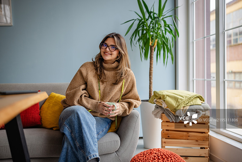年轻女性在舒适的客厅中放松，手中拿着一杯，周围是五彩缤纷的靠垫和植物。图片素材