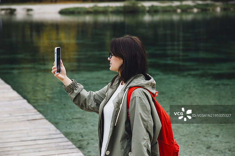 侧面视角的女性穿着雨衣，站在湖边用手机拍照图片素材