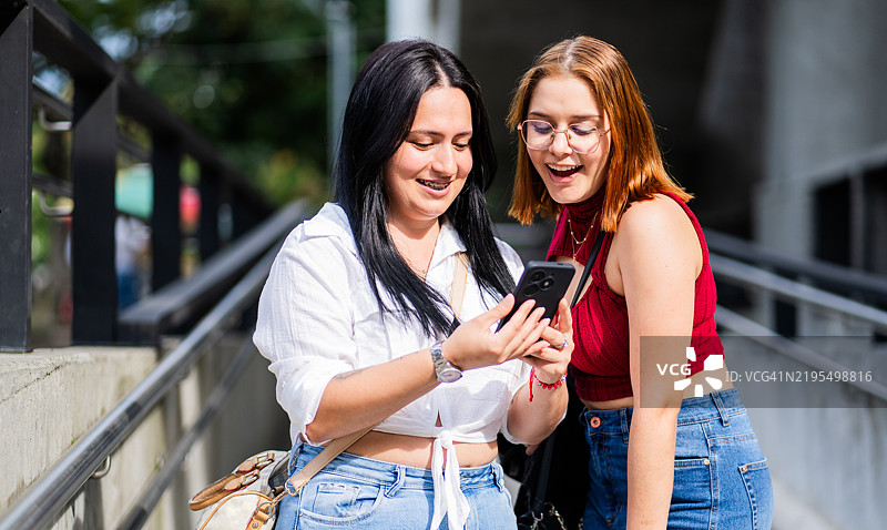 两位年轻女性在街上微笑并查看手机的肖像。图片素材