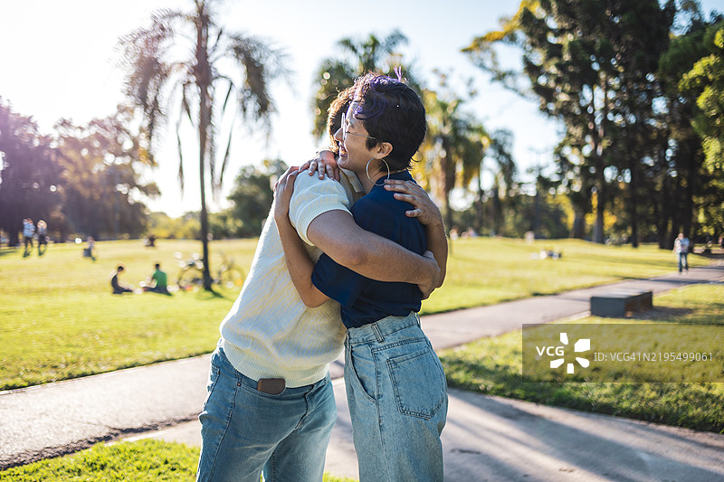 两个朋友在阳光明媚的公园里拥抱，周围是树木和绿意。图片素材