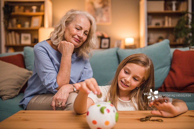 一个无忧无虑的小女孩在客厅里和她的奶奶一起，把硬币放进储蓄罐。图片素材