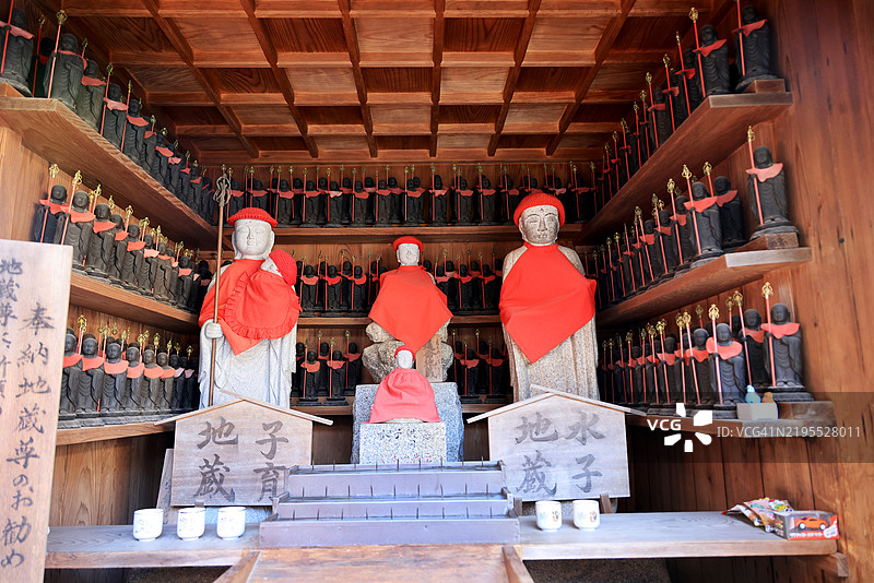 在日本宫岛的大愿寺，三尊大型地藏菩萨雕像穿着红色围兜和帽子，周围环绕着许多小型地藏菩萨雕像，角落里放着玩具供品。图片素材