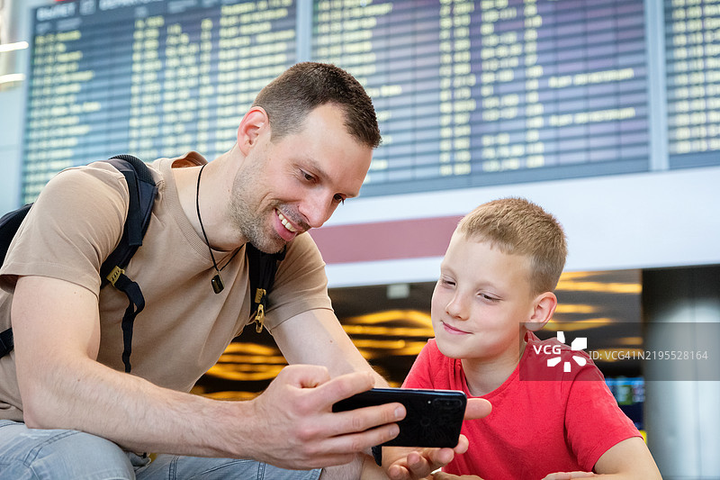 移动游戏让年轻男孩和爸爸在出发显示屏下团聚。机场等待变成了与智能手机娱乐的高质量亲子时光。图片素材