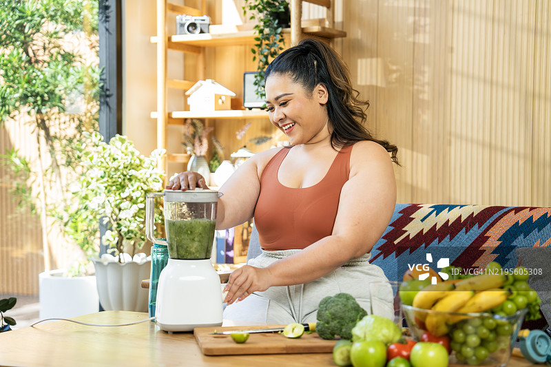 微笑的女性在明亮的家居环境中准备健康的奶昔，体型丰满。图片素材