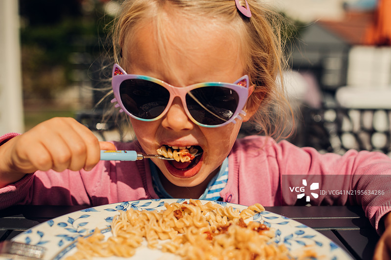 小女孩在户外戴着太阳镜吃意大利面配番茄酱。图片素材