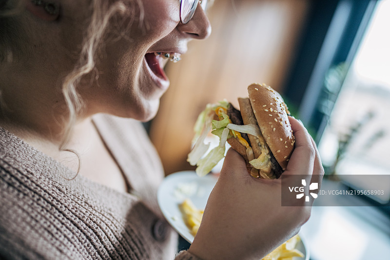 厨房里一位快乐女性的特写，她手里拿着一盘舒适食物，正咬一口汉堡。图片素材