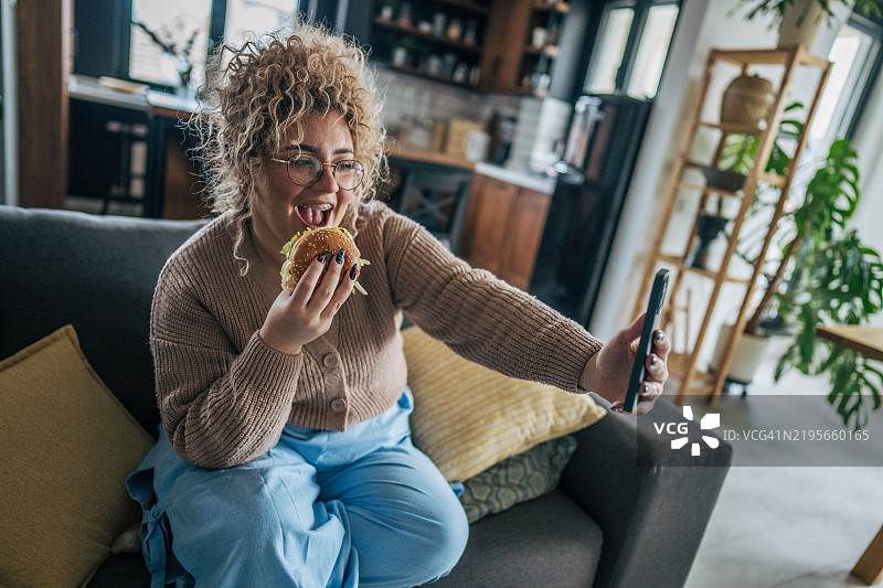 快乐的女人在沙发上享受汉堡，同时自拍图片素材