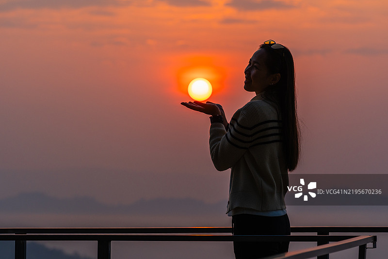 一位女性顽皮地摆姿势，仿佛用双手托起升起的太阳，背对着雾气缭绕的山脉，映衬着绚丽的日出。图片素材