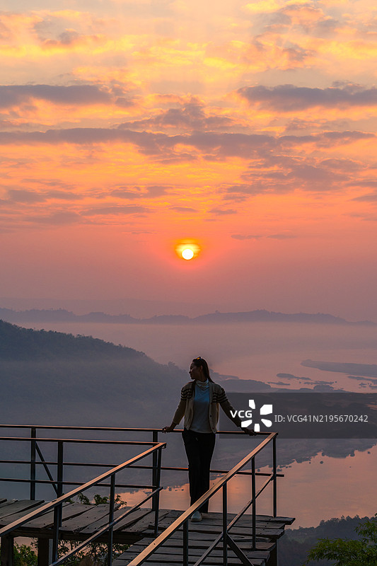 一位女性站在观景平台上，拍摄美丽的日出，背景是雾蒙蒙的山脉和宁静的河谷。图片素材