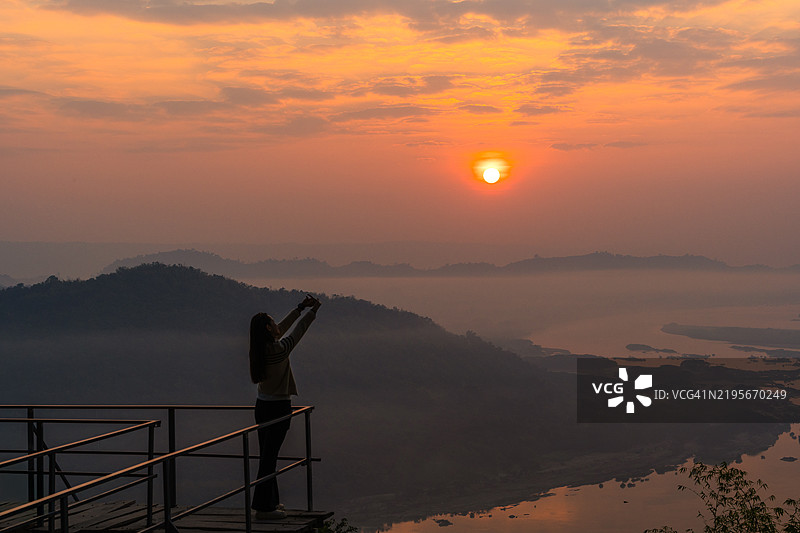 一位女性站在观景平台上，拍摄美丽的日出，背景是雾蒙蒙的山脉和宁静的河谷。图片素材