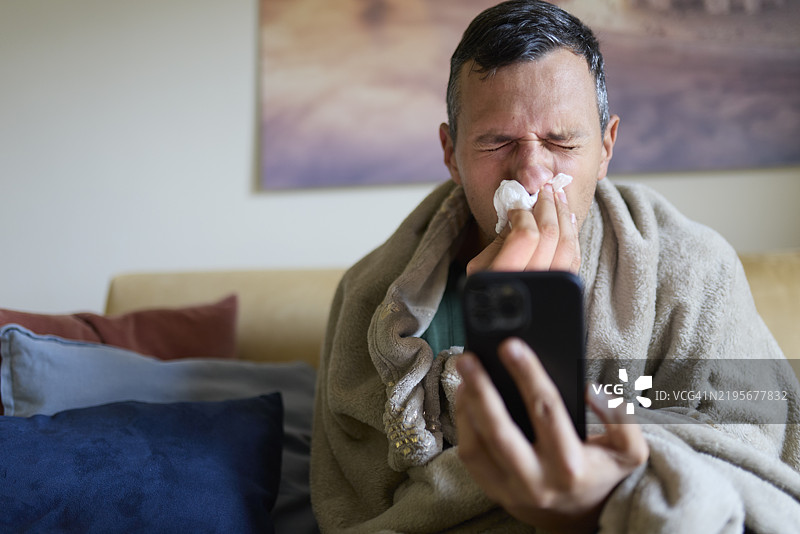 生病的男人坐在沙发上使用智能手机。图片素材