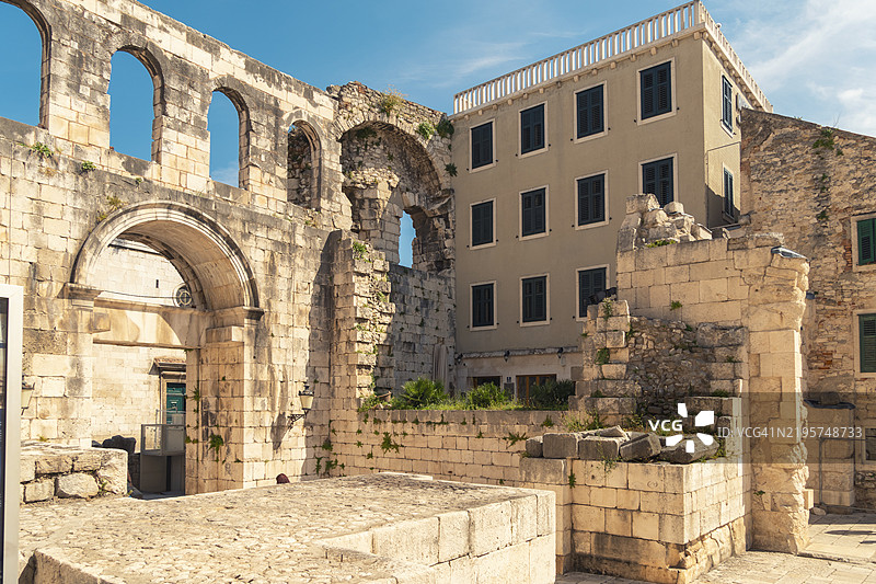 古老的石拱门矗立在现代建筑旁，蓝天清澈，历史建筑与当代建筑在克罗地亚斯普利特的海伦娜女王草地交融。图片素材