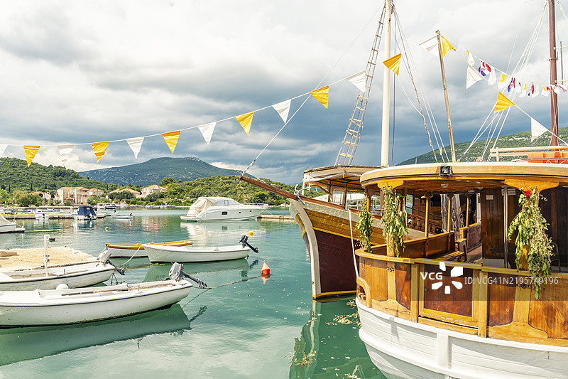 五彩斑斓的旗帜在宁静的港口上空飘扬，船只停泊在此，背景是云雾缭绕的山脉，地点：克罗地亚马里斯通。图片素材