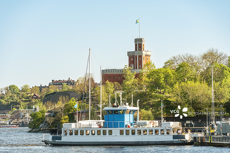 名为Djurgarden 9的客轮在晴朗的日子里停靠在郁郁葱葱的绿树和历史悠久的斯德哥尔摩城堡前，瑞典斯德哥尔摩。图片素材