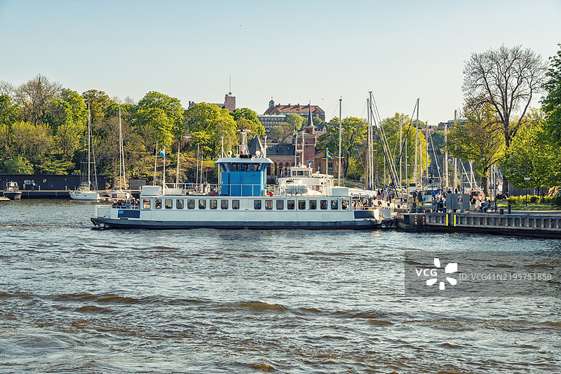 在瑞典斯德哥尔摩的斯凯普霍尔门，一个阳光明媚的日子里，停靠在码头的蓝白色渡轮，背景中有行人和帆船。图片素材