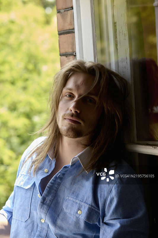 长发男子靠在窗台上，神情若有所思。图片素材