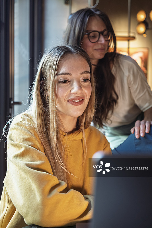 微笑的女性穿着黄色连帽衫，正在用笔记本电脑工作，身后站着另一位女性，德国柏林图片素材