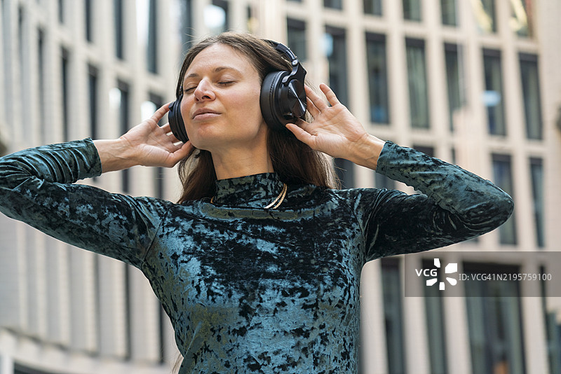 穿着蓝色天鹅绒上衣的女性闭着眼睛，享受着耳机中的音乐，站在现代建筑前，德国柏林。图片素材