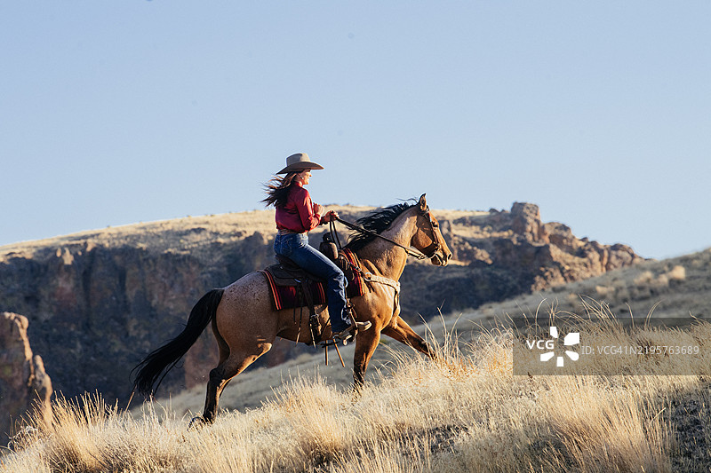 一位戴着宽边帽、穿着红色衬衫的女性骑着马在阳光明媚的日子里沿着草坡向上骑行，背景是爱达荷州的岩石景观。图片素材