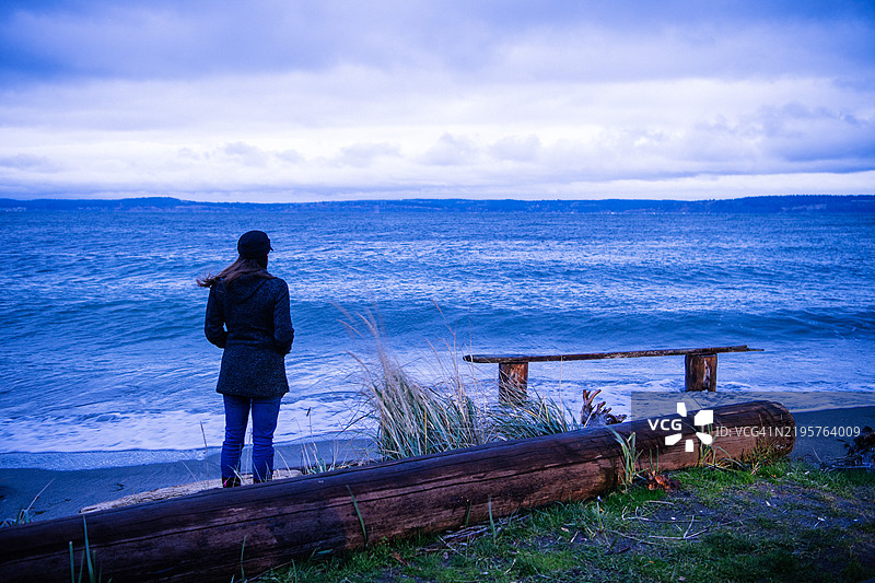 一位女性站在草坪海岸线上，背对着镜头，穿着深色外套和牛仔裤，地点：华盛顿州挪威角，美国。图片素材