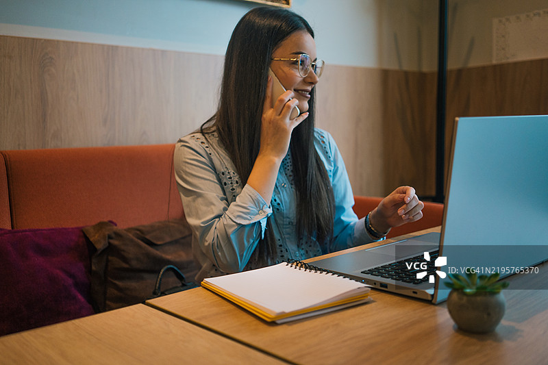 一位年轻的女商人，穿着柔和的蓝色衬衫，佩戴着时尚的眼镜，坐在咖啡馆温馨的氛围中。她面带微笑，轻松地进行电话交谈，同时在笔记本电脑上工作。图片素材