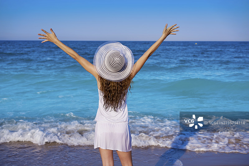 穿着夏季白色连衣裙、在海中张开双臂的女孩戴着海滩帽图片素材