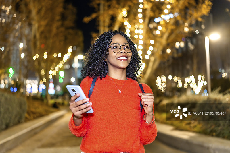 微笑的学生在夜晚的美丽装饰城市中走着，使用智能手机与朋友联系并导航节日街道。图片素材