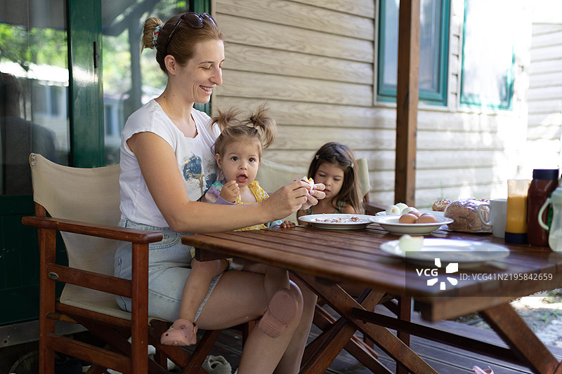 一位快乐的女人坐在门廊上，和她的女儿们一起剥鸡蛋准备早餐。图片素材