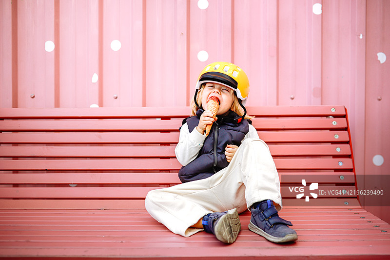 一个可爱的男孩戴着黄色头盔和背心，在户外咖啡馆的粉色长椅上享受冰淇淋，创造有趣的童年回忆。图片素材