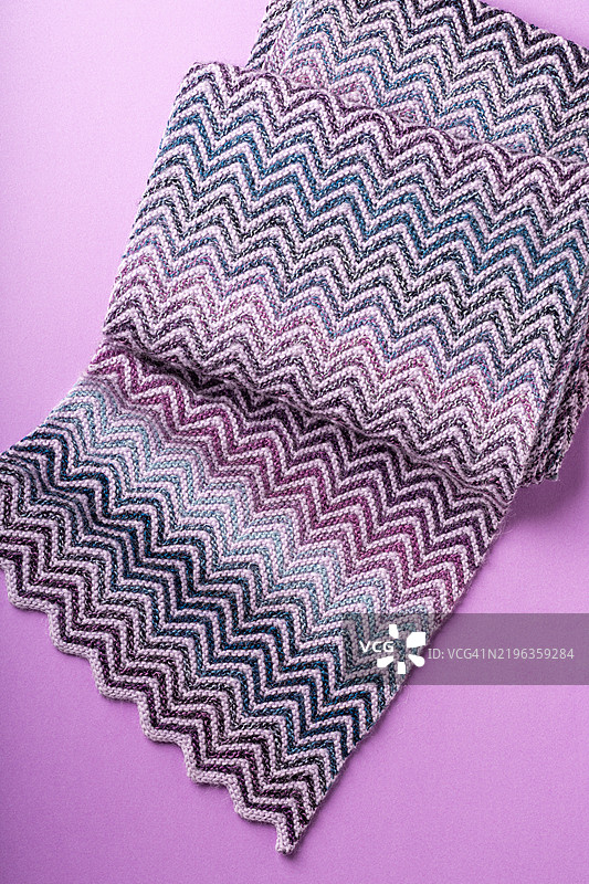 柔软的针织围巾特写，呈现紫色、蓝色和灰色的锯齿形图案，背景为淡紫色。舒适而时尚的冬季配饰。图片素材