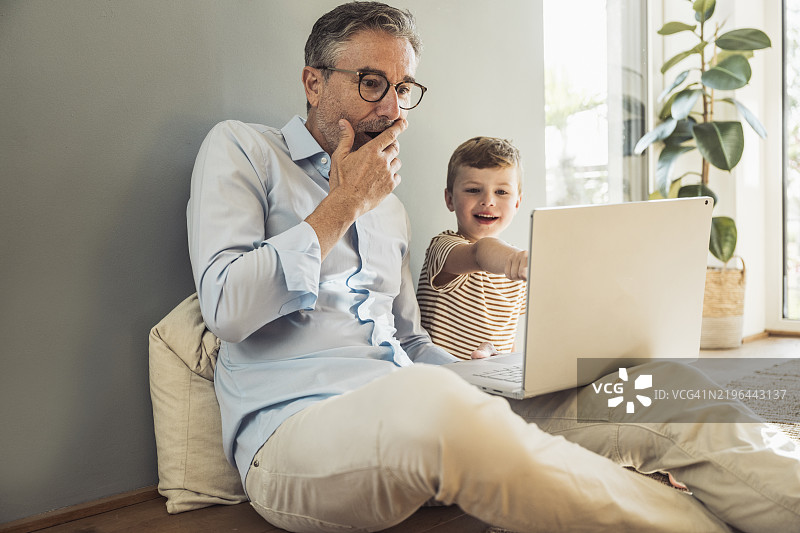 在家中，孙子旁边的男人用手捂嘴，正在使用笔记本电脑。图片素材