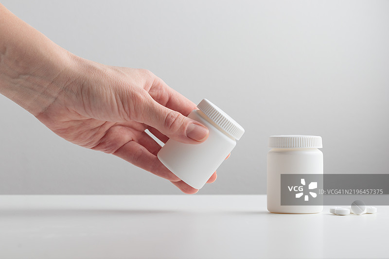 一位年轻女性的手在白色背景上拿着一个白色塑料瓶，药品、胶囊、补充剂或维生素的品牌包装模版，健康护理概念。图片素材