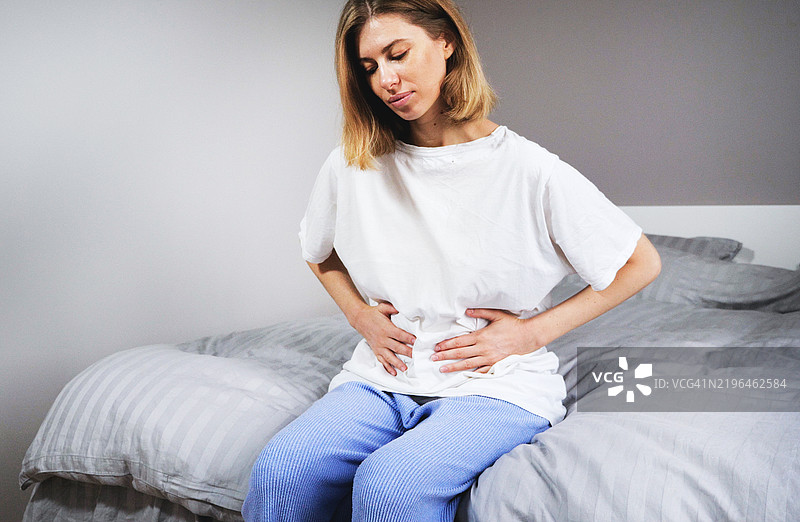 年轻的金发女性坐在家里的沙发上，感到腹部疼痛。图片素材