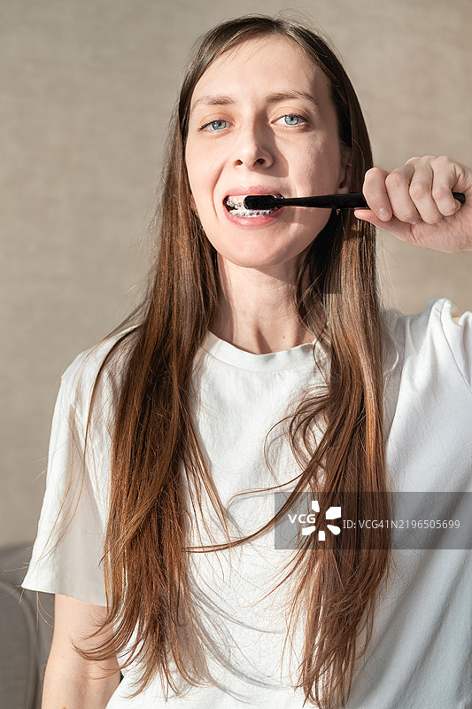 一位穿着白色T恤、长发的年轻女性在白色背景下用牙刷刷牙，牙齿上戴着牙套。图片素材