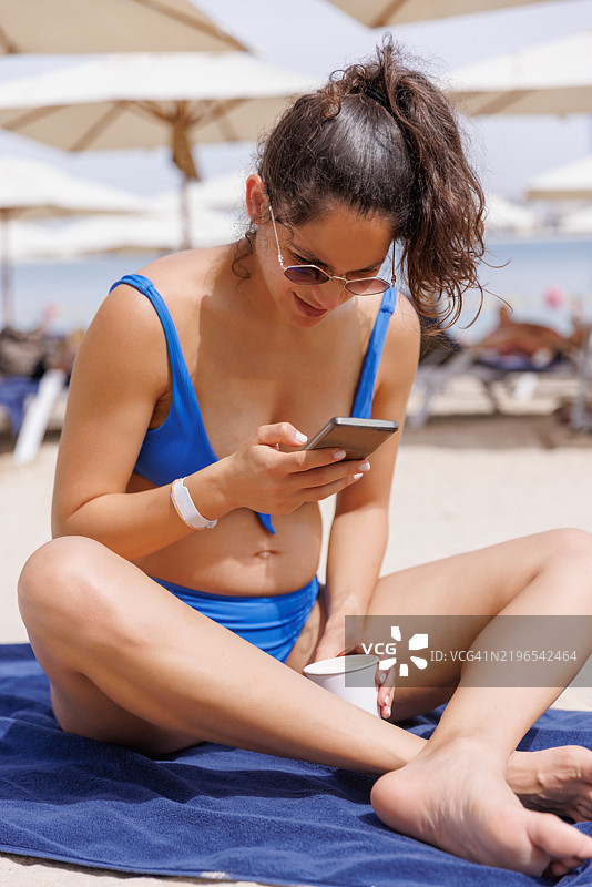 美丽的孕妇穿着蓝色比基尼，坐在沙滩毛巾上用智能手机发信息图片素材