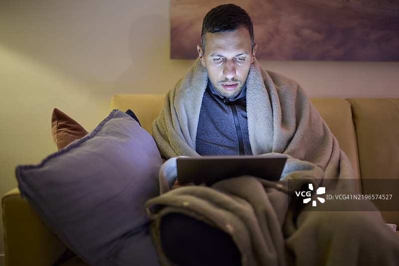 生病的男人晚上坐在沙发上使用平板电脑。图片素材