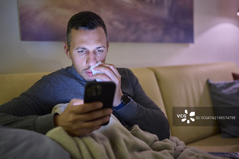 生病的男人晚上在家沙发上使用智能手机图片素材