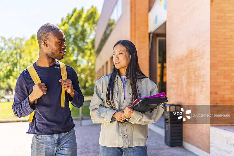 两位非洲和中国的多民族朋友在大学校园里边走边谈，手里拿着文件夹和笔记本。图片素材