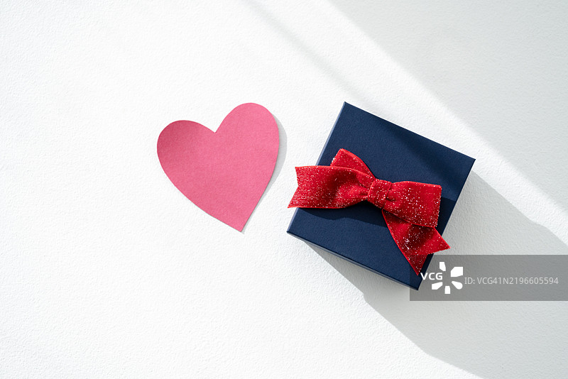 白色表面上带有红色蝴蝶结和心形切口的礼品盒，阳光明媚。图片素材