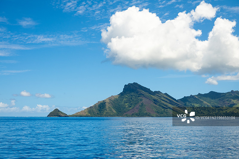 一座热带岛屿被棕榈树和茂密的植被环绕，从船上望去，四周是明亮阳光下的生动碧蓝海水。图片素材
